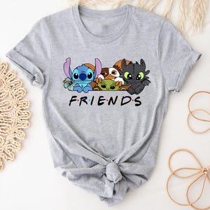 May include: A light gray t-shirt featuring a graphic of cartoon characters, including Stitch, Baby Yoda, and Toothless, above the word "FRIENDS" in a familiar font. The shirt is knotted at the bottom.