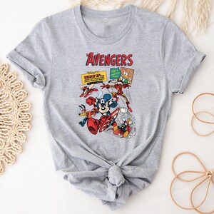 May include: A grey t-shirt with a vibrant graphic of Disney characters in an Avengers theme. The design showcases Mickey Mouse as Captain America, Donald Duck, and Iron Man, with the word "Avengers" in red at the top.