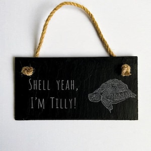May include: Black slate sign with a rope hanger. The sign has a white drawing of a turtle and the text "Shell yeah, I'm Tilly!"