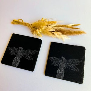 May include: Two black slate coasters with a white line drawing of a fairy with wings outstretched. The coasters are on a white surface with a bouquet of dried flowers.