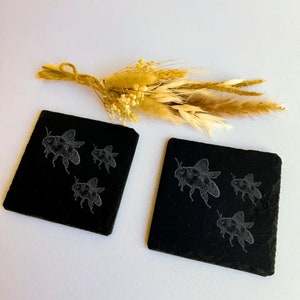 May include: Two black slate coasters with a white etched design of bees. The coasters are square and have rounded corners. The bees are in flight and have detailed wings.