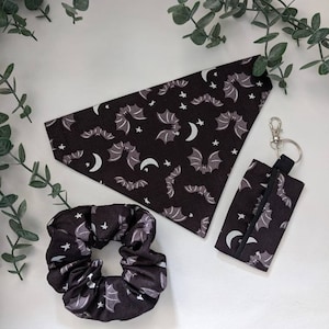 Könnte beinhalten: Ein schwarzes und graues Accessoire-Set zum Thema Fledermaus. Das Set enthält ein dreieckiges Halstuch, ein Scrunchie und eine kleine Tasche mit einem silberfarbenen Clip. Die Artikel weisen ein Muster aus Fledermäusen, Sternen und Monden auf.