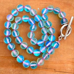 May include: A necklace featuring iridescent, round beads in shades of blue and green, strung on pink thread. The necklace has a silver-colored toggle clasp. The beads have a subtle, glowing effect.