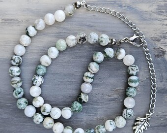 May include: A beaded necklace featuring round white and green mottled stone beads arranged in a circular pattern. The necklace includes a silver-toned metal chain extender with a lobster claw clasp and a small silver-toned leaf-shaped charm.