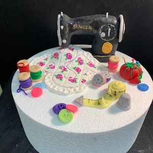 May include: A white cake decorated with a black sewing machine, a white fabric with pink flowers, spools of thread, buttons, a measuring tape, and a thimble.