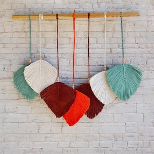 May include: Six macrame feather wall hangings in various colors, including white, green, red, and orange. The feathers are hanging from a wooden dowel rod.