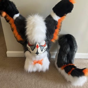 May include: A furry costume head and tail set. The head is primarily white, gray, and black, with orange accents and a stylized face. The tail mirrors the color scheme. The costume is designed to resemble a stylized animal.