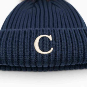 Könnte beinhalten: Marineblaue Strickmütze mit weiß gesticktem "C" auf der Vorderseite. Die Mütze hat eine gerippte Struktur und einen flauschigen Bommel oben. Ein Winteraccessoire.