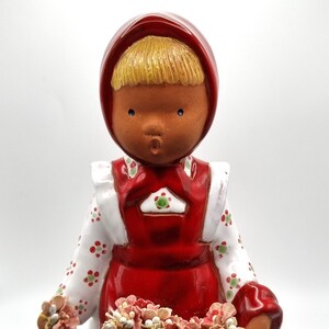 May include: A ceramic figurine of a girl wearing a red dress and a red bonnet. She is holding a basket of flowers in her hands.