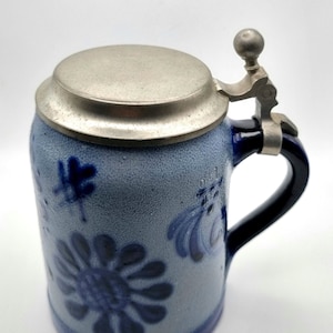 May include: A blue and white ceramic beer stein with a pewter lid and a blue handle. The stein features a floral design and the number "051" is visible on the side.