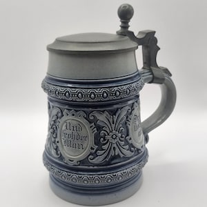 May include: A blue and grey ceramic beer stein with a hinged lid and handle. The stein features embossed floral designs and the words "Und frohder Mann" in an oval. A traditional drinking vessel.