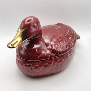 May include: A red ceramic duck-shaped serving dish with a gold-painted beak and accents. The dish has a textured surface that resembles feathers.