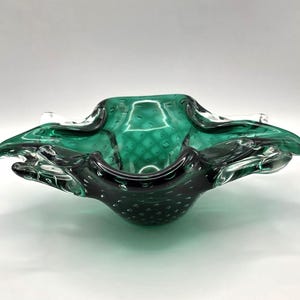 May include: An emerald green glass bowl with an unusual, organic form. The bowl has a textured surface with tiny air bubbles and a clear, ruffled edge. This decorative piece is perfect for displaying small items, adding a touch of elegance.