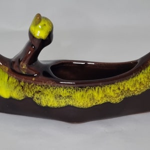 May include: A brown ceramic canoe planter with a yellow glaze. The canoe has a small figure of a person paddling in the front.