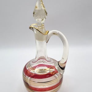 May include: A clear glass decanter with a handle and a stopper. The base of the decanter has red and gold horizontal stripes. The stopper and rim are trimmed with gold. The handle is made of clear glass.