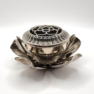 May include: A silver incense burner, shaped like a lotus flower. The burner features a detailed, openwork lid and rests within the flower's petals. The design is intricate and ornate, with a focus on floral elements.