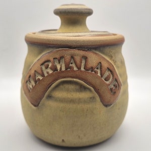 May include: A ceramic jar with a lid, decorated with a brown and white glaze. The jar has the word "MARMALADE" written on it in a brown glaze.