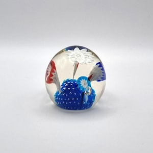 May include: A clear glass paperweight with a blue, white, and red floral design. The paperweight has a round shape and features a blue base with white and red accents, and a white flower.