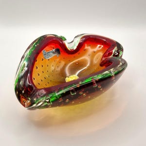 May include: A vintage Murano glass ashtray. The ashtray has a striking colour scheme of red, green, and amber. The glass is textured with small bubbles throughout. A small label is visible on the side.