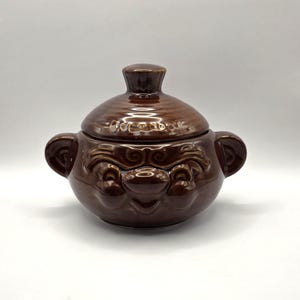 May include: A brown glazed ceramic lidded pot featuring a sculpted face with prominent eyebrows, a round nose, and two side handles. The lid has a matching brown finish and a central knob handle, creating a cohesive look for this kitchen vessel.