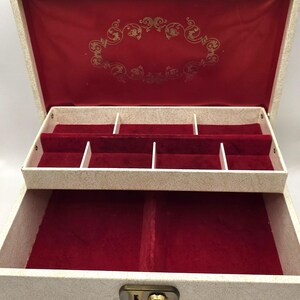 May include: Cream-coloured jewellery box with a red velvet interior. The box has multiple compartments and a gold-coloured clasp. The inside lid features a gold decorative design. The box is designed to store jewellery.