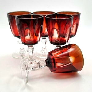 May include: Set of six red wine glasses with a clear glass stem and base. The glasses have a faceted design and are arranged in a group on a white surface.