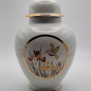 May include: White porcelain jar with a gold lid and a gold rim. The jar features a circular design depicting a hummingbird in flight, surrounded by yellow and brown irises.