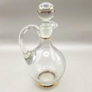 May include: Clear glass decanter with a round body, handle, and spout. The stopper and base are accented with gold bands. The decanter has a decorative etched floral design and is suitable for serving beverages.