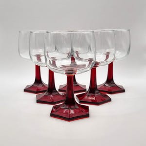 May include: Six clear wine glasses with ruby red stems and bases. The glasses are arranged in a semi-circle, showcasing their unique design. The stems have a geometric shape, adding a touch of elegance to the glassware.