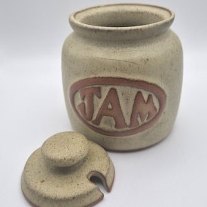 May include: A beige ceramic jam jar with a lid and a brown "JAM" inscription.