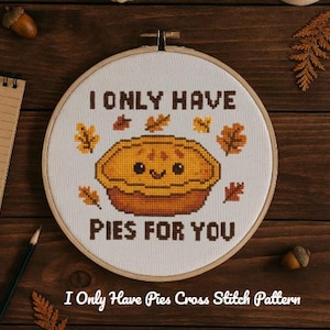 Può includere: Un motivo a punto croce in un telaio di legno presenta una torta sorridente con la scritta "I ONLY HAVE PIES FOR YOU". Foglie autunnali circondano la torta. Il testo "I Only Have Pies Cross Stitch Pattern" è in basso.