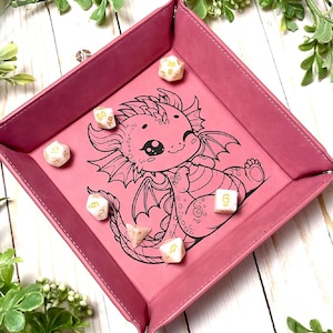 May include: A pink felt dice tray with a black and white illustration of a cute dragon. The tray has a raised edge to keep dice from rolling off. There are several white dice with gold numbers inside the tray.
