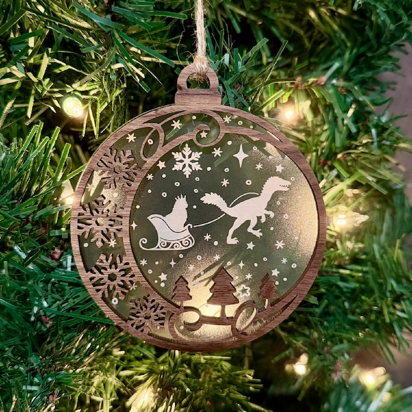 May include: A circular wooden Christmas ornament featuring a laser-cut frame with snowflake patterns. The center displays a white silhouette of a dinosaur pulling a sleigh with a cat inside, set against a green background with stars.