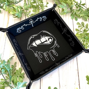 May include: Black leather dice tray with a white design of a dripping vampire mouth with fangs. The tray has a stitched border with a repeating design of a cross and a swirl.