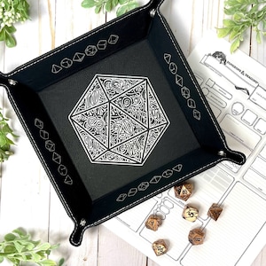 May include: Black leather dice tray with a white, intricate design of a 20-sided die. The tray has a stitched border and is shown with a set of dice on a white Dungeons & Dragons character sheet.