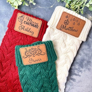 May include: Three green, red, and white knitted stockings with brown leather tags. The tags have personalized names and festive designs, including a dice, a Christmas tree, and a dragon.