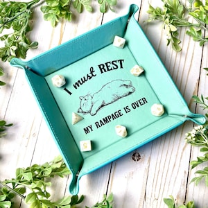 May include: A turquoise dice tray with a cat illustration and the text "I must rest, my rampage is over". The tray has six white dice with gold numbers.