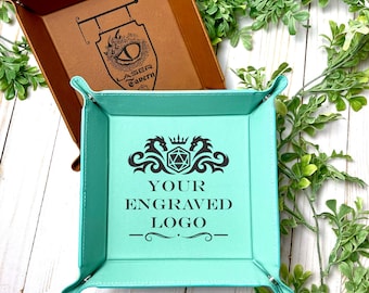 May include: A square mint green valet tray with snap corners and a black graphic featuring a crown, dragons, and a twenty-sided die above the text YOUR ENGRAVED LOGO, positioned in front of a brown valet tray with a LASER TAVERN logo.