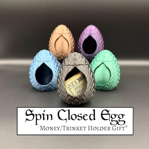 May include: Five colorful, egg-shaped trinket holders. The holders are in shades of blue, bronze, purple, gray, and green. One gray holder contains a twenty-dollar bill. The text "Spin Closed Egg MONEY/TRINKET HOLDER GIFT*" is at the bottom.