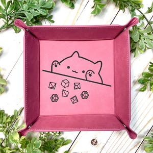 May include: Pink felt dice tray with a black outline of a cat peeking over the edge and looking at dice. The cat has a surprised expression. The dice are scattered on the tray.