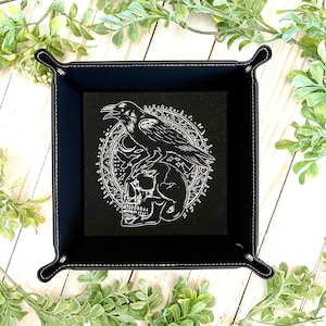 May include: Black leather dice tray with a white design of a raven perched on a skull surrounded by a circular pattern.