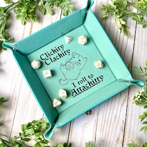 May include: A turquoise felt dice tray with a white cat illustration and the text "Clickity Clackity I roll to Attackitty". The tray has six white dice with black numbers.