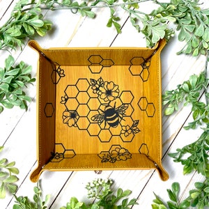 May include: A small, square, yellow leather tray with a black bee and honeycomb design. The tray has a stitched edge and is perfect for holding small items.
