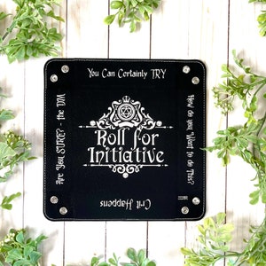 May include: Black leather dice tray with white stitching and silver rivets. The tray features a white graphic with the text "Roll for Initiative" and a crown with a dice symbol. The text "You Can Certainly TRY" and "How do you Want to do This?" are on the top and side of the tray. The text "Crit Happens" is on the bottom of the tray.