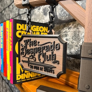 May include: A wooden sign for "The Desperado Club" with a sword graphic, hanging from a chain. The sign is brown with black lettering and trim. Colorful books with the text "Dungeon Crawler" are visible to the left.