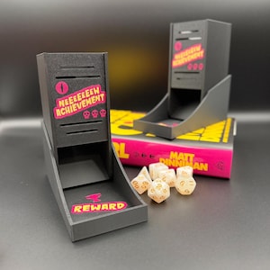 May include: Black dice tower with pink text that reads "NEEEEEEEW ACHIEVEMENT" and "REWARD". The tower has a tray to catch dice. Six off-white dice with gold numbers are next to the tower. The tower is sitting on a book.