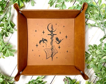 May include: A brown leather dice tray with a design of a sword, a book, and three dragons in a starry sky.