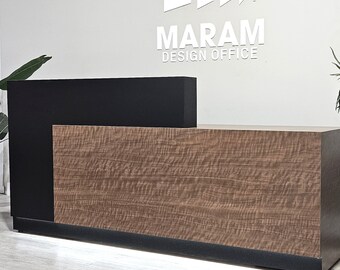 May include: A modern reception desk with a black and wood laminate finish. The desk has a black countertop and a lower wood panel. The desk is lit with a strip of LED lights. The desk is in front of a white wall with the text "MARAM DESIGN OFFICE" in white letters.
