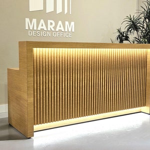 May include: A wooden reception desk with a slatted front panel and a light strip behind the slats. The desk is lit from below. The text "MARAM DESIGN OFFICE" is on the wall behind the desk.