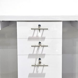 May include: White office desk with three drawers. Each drawer has a silver handle and a keyhole. The desk is on a gray floor.